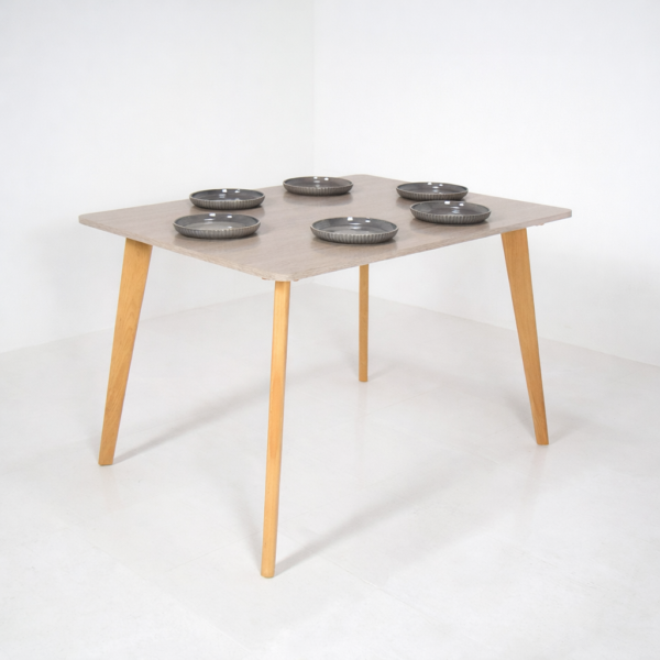 Table à manger carrée design scandinave avec plateau clair et pieds en bois naturel, présentée sur fond blanc avec vaisselle.
